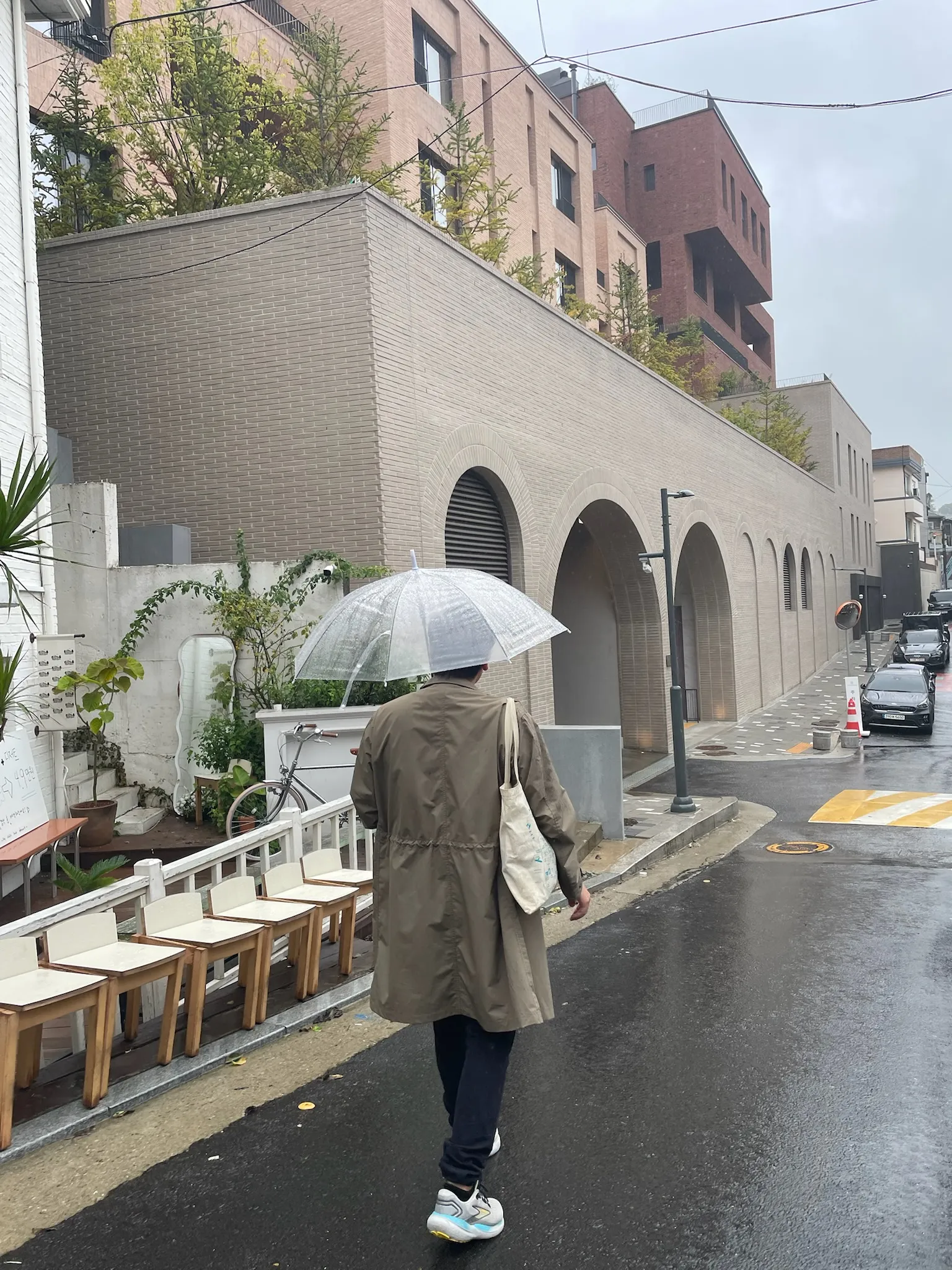 Quinn wearing a beige trench coat and navy pants holds a clear umbrella while walking on a wet street past a modern brick building with large arched openings