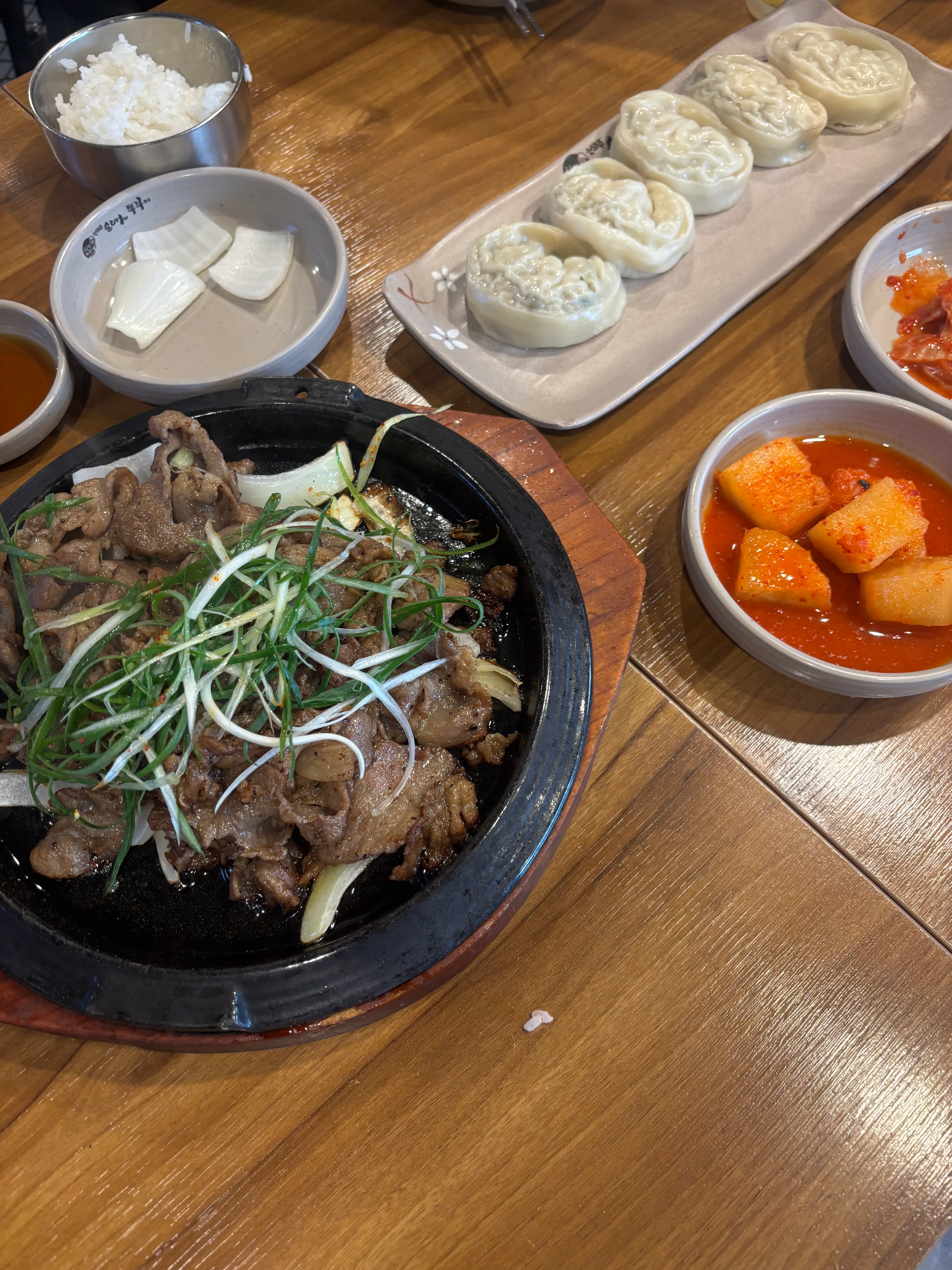Table spread of Korean food including a black stone bowl of beef and vegetables garnished with green onions, steamed dumplings filled with meat on a rectangular plate, and a metal bowl of white rice