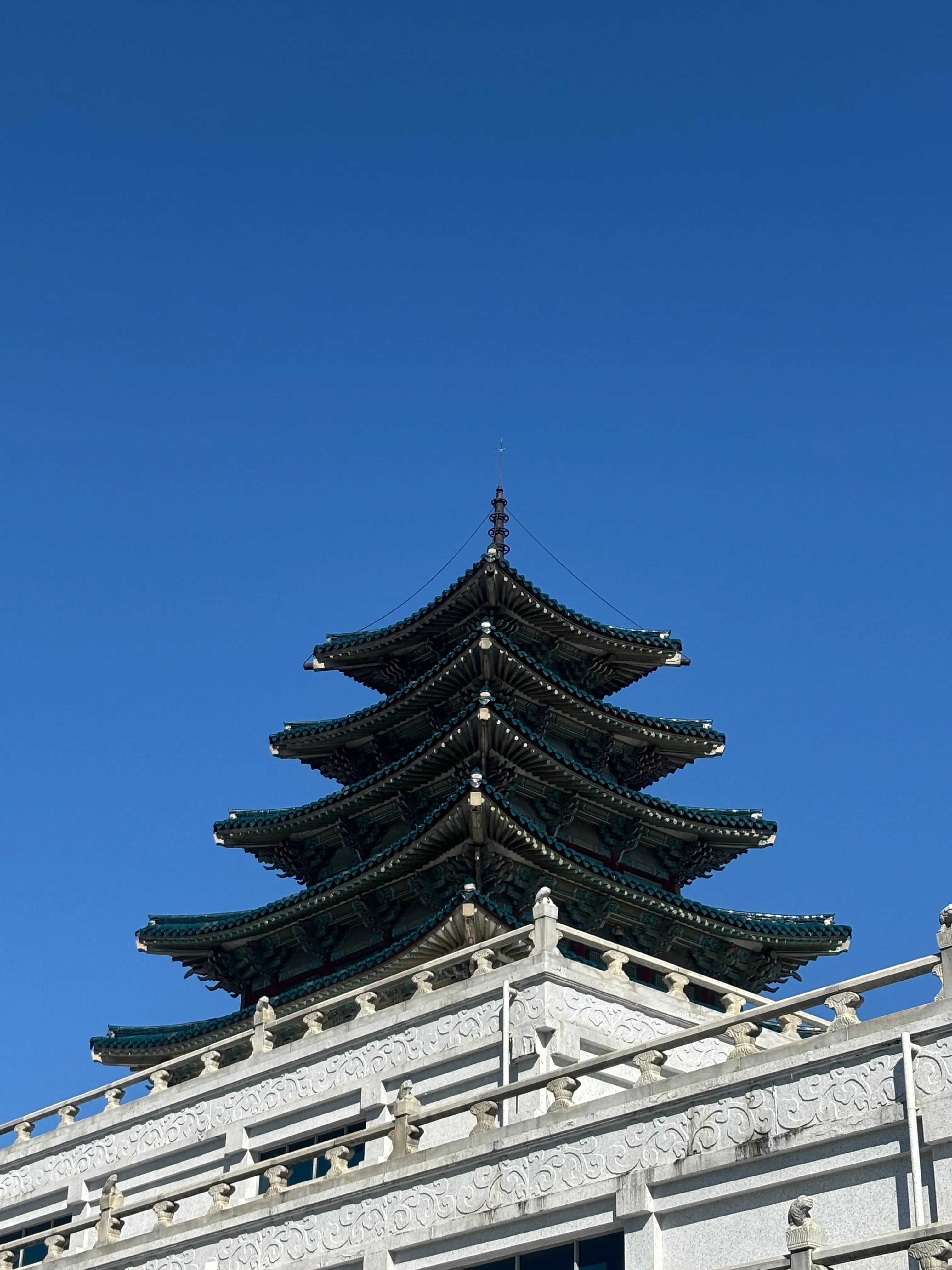 An ornate East Asian pagoda with five tiers and curved, upturned eaves topped with green glazed tiles and a spire, featuring white decorative railings with carved patterns on the tier below, set against a clear blue sky