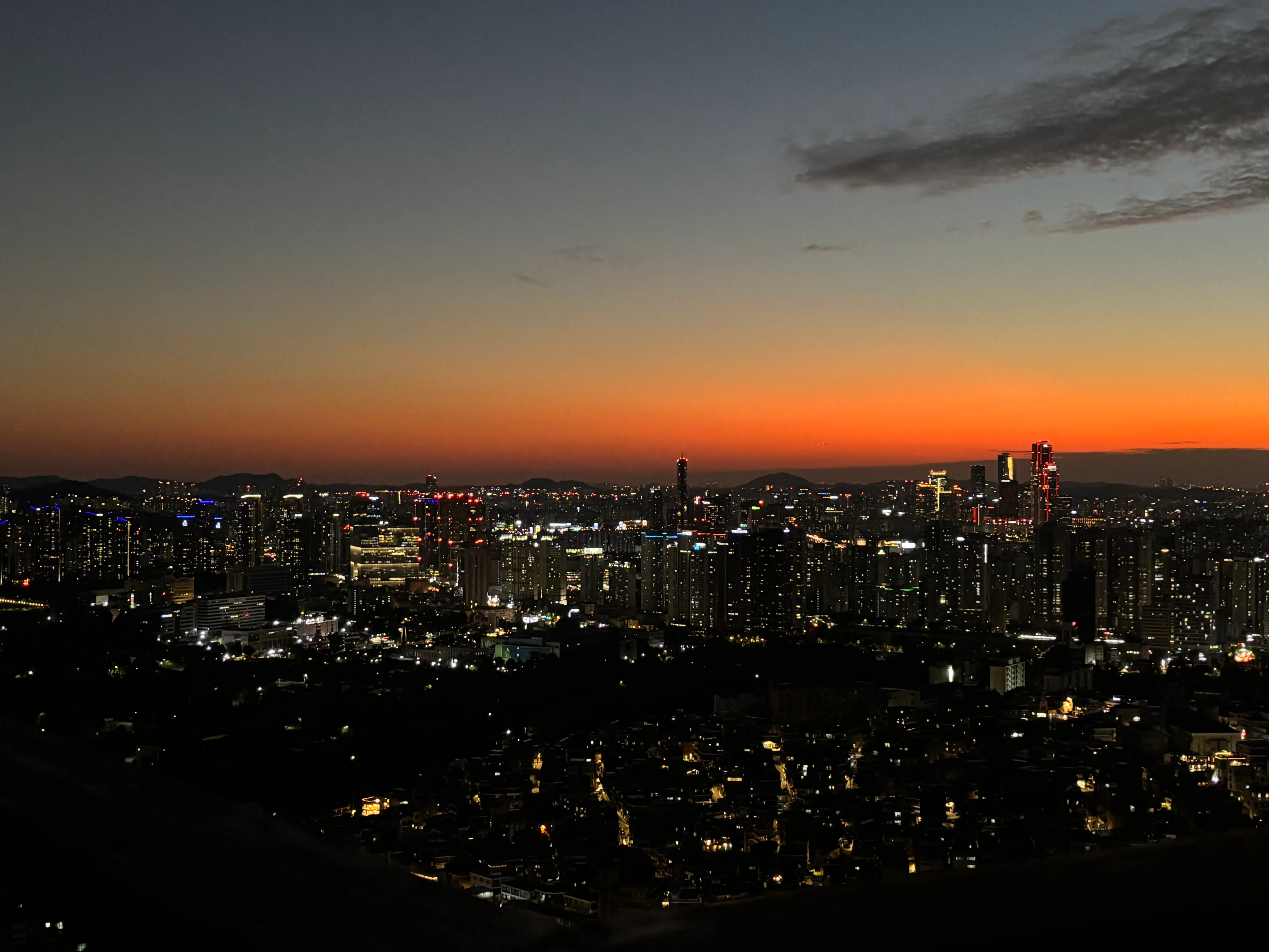 Hundreds of buildings with twinkling lights under a deep orange sunset, viewed from the a road leading toward Namsan Seoul Tower