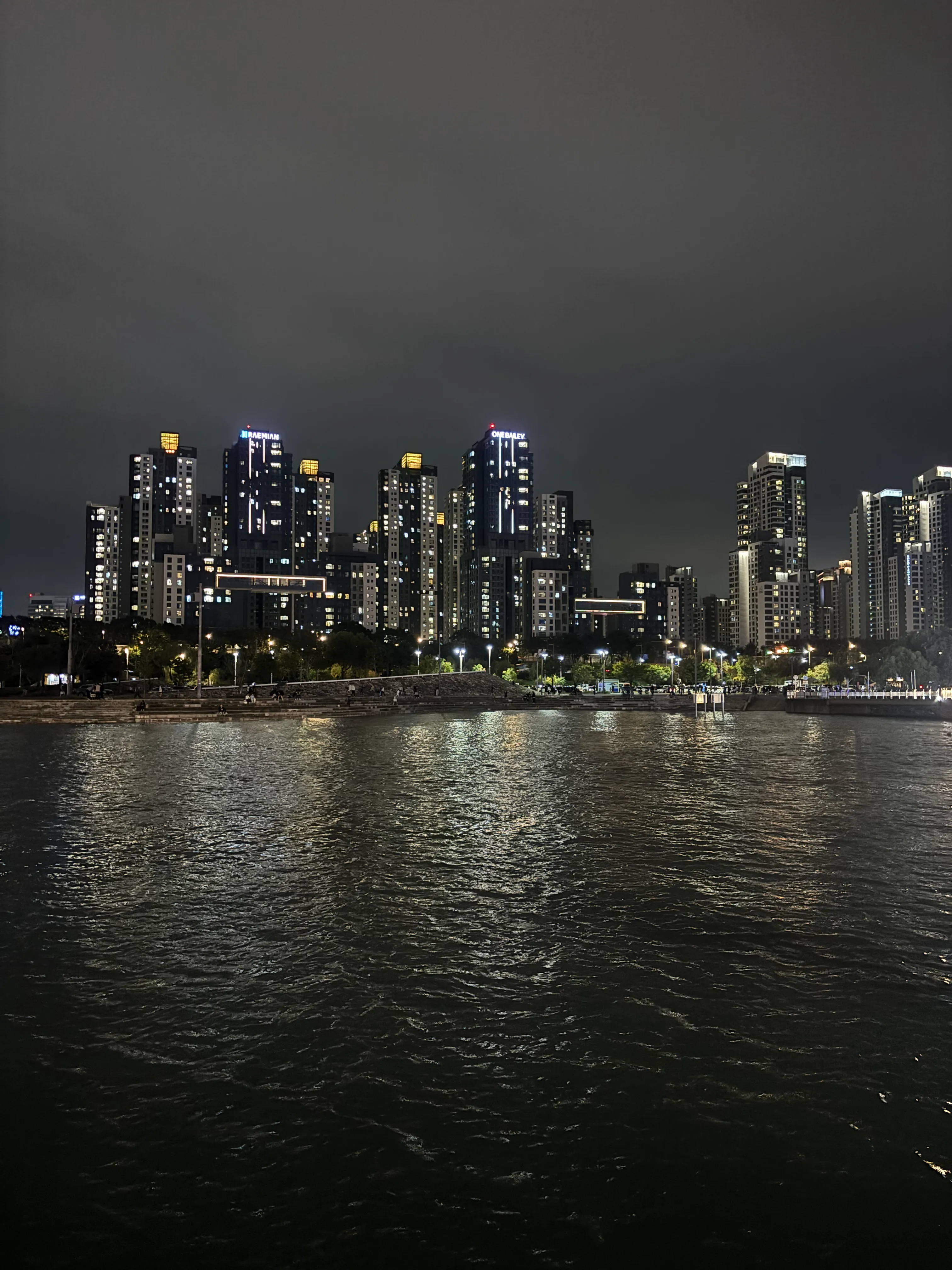 A nighttime cityscape view across a body of water showing an illuminated high-rise skyline with numerous tall residential and commercial buildings lit up against a dark gray sky, with the Han river below