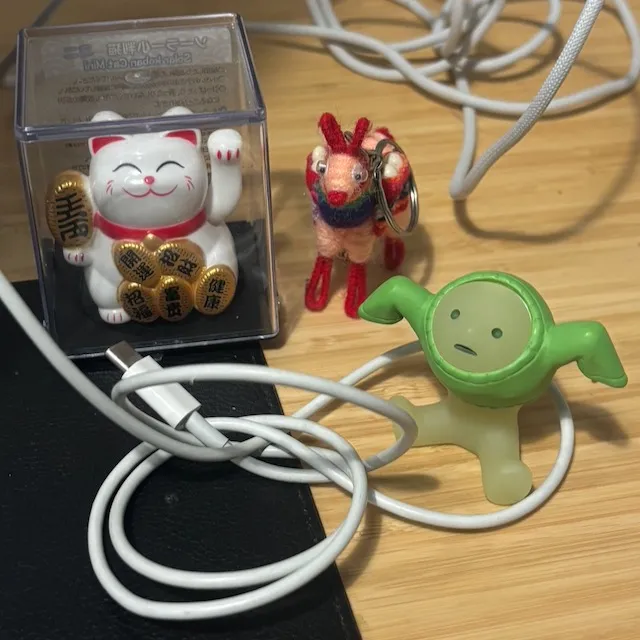 Waving Maneki-neko cat, small figurine of a person putting on a sweater, and a felt and wire alpaca keychain, all surrounded by jumbled cables