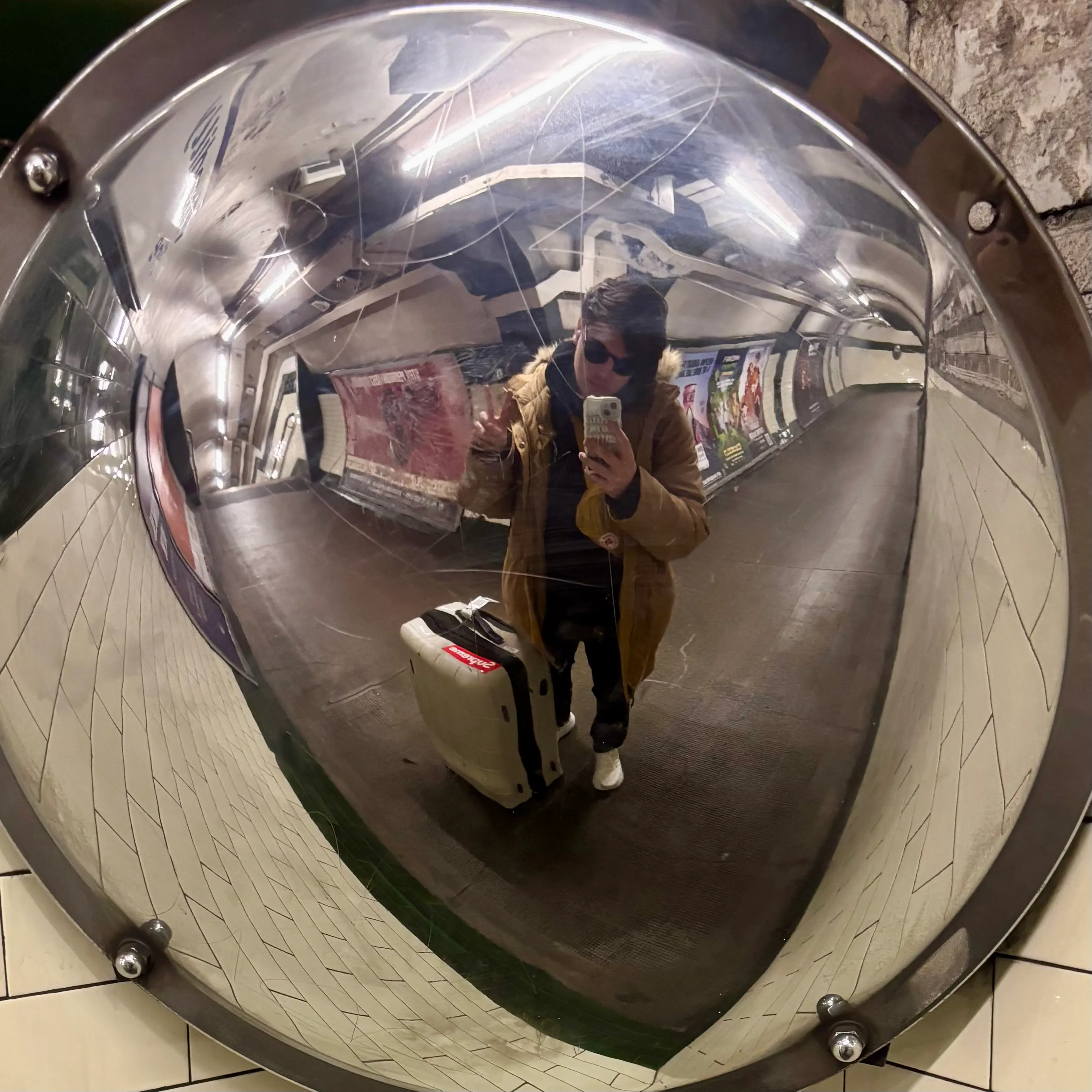 Quinn, wearing sunglasses, holding a peace sign, standing in front of a fish-eye mirror at the corner of intersecting hallways in a London Underground station.