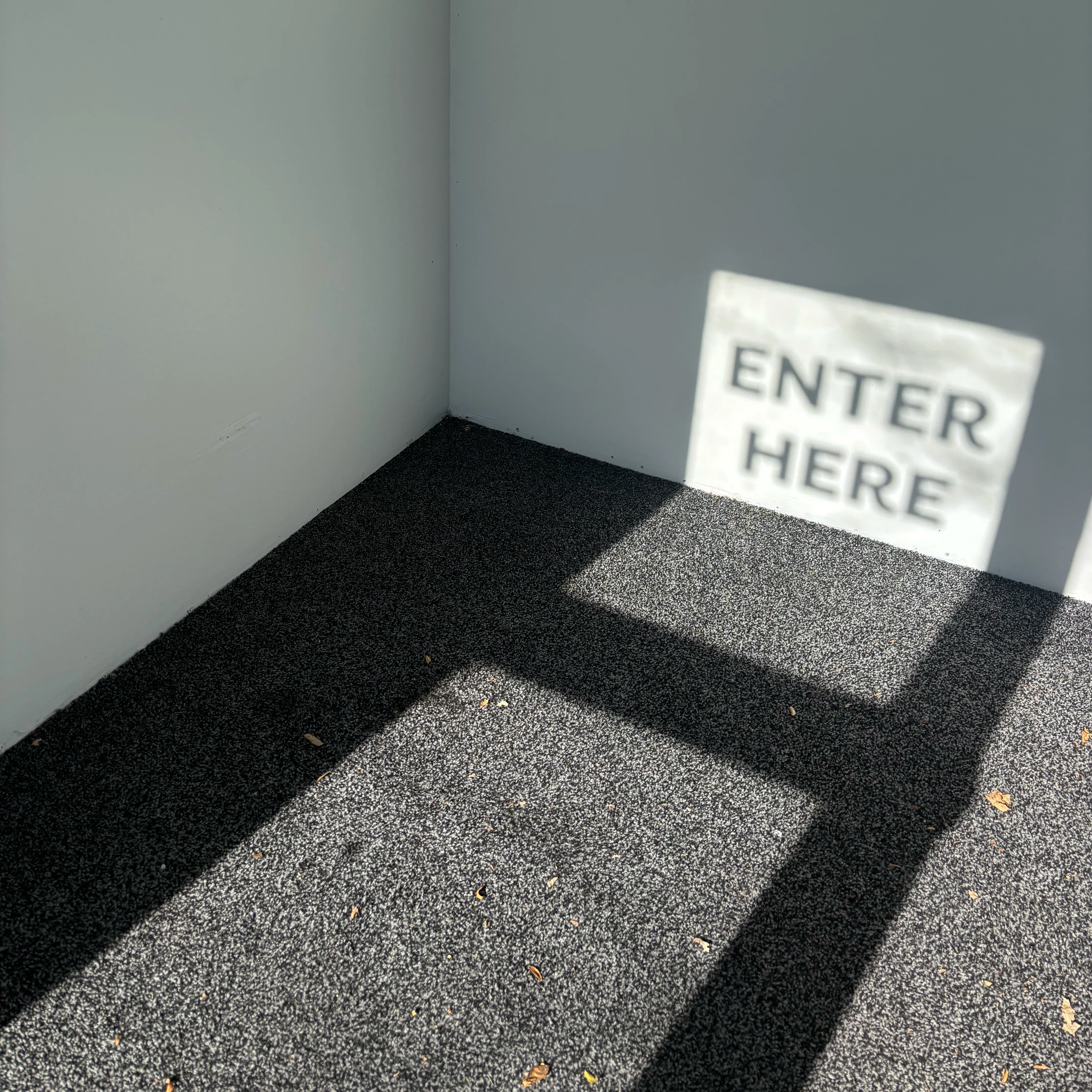 Corner of a building with a black textured doormat and the shadow of an 'ENTER HERE' sign against a grey wall