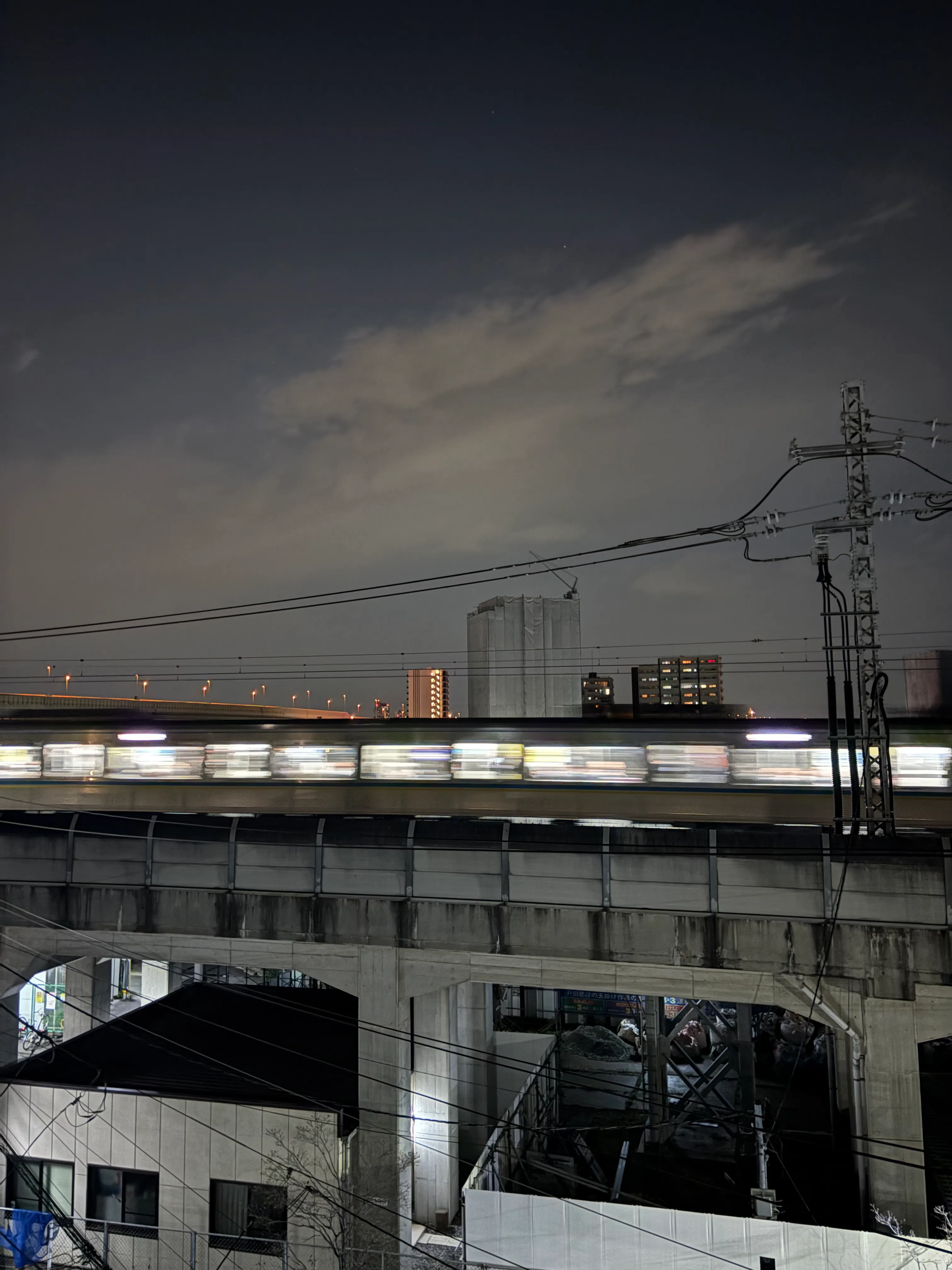 A train passing on an elevated track at dusk, captured with motion blur showing horizontal streaks of light from the train windows. Power lines and urban buildings are visible in the background against a darkening sky