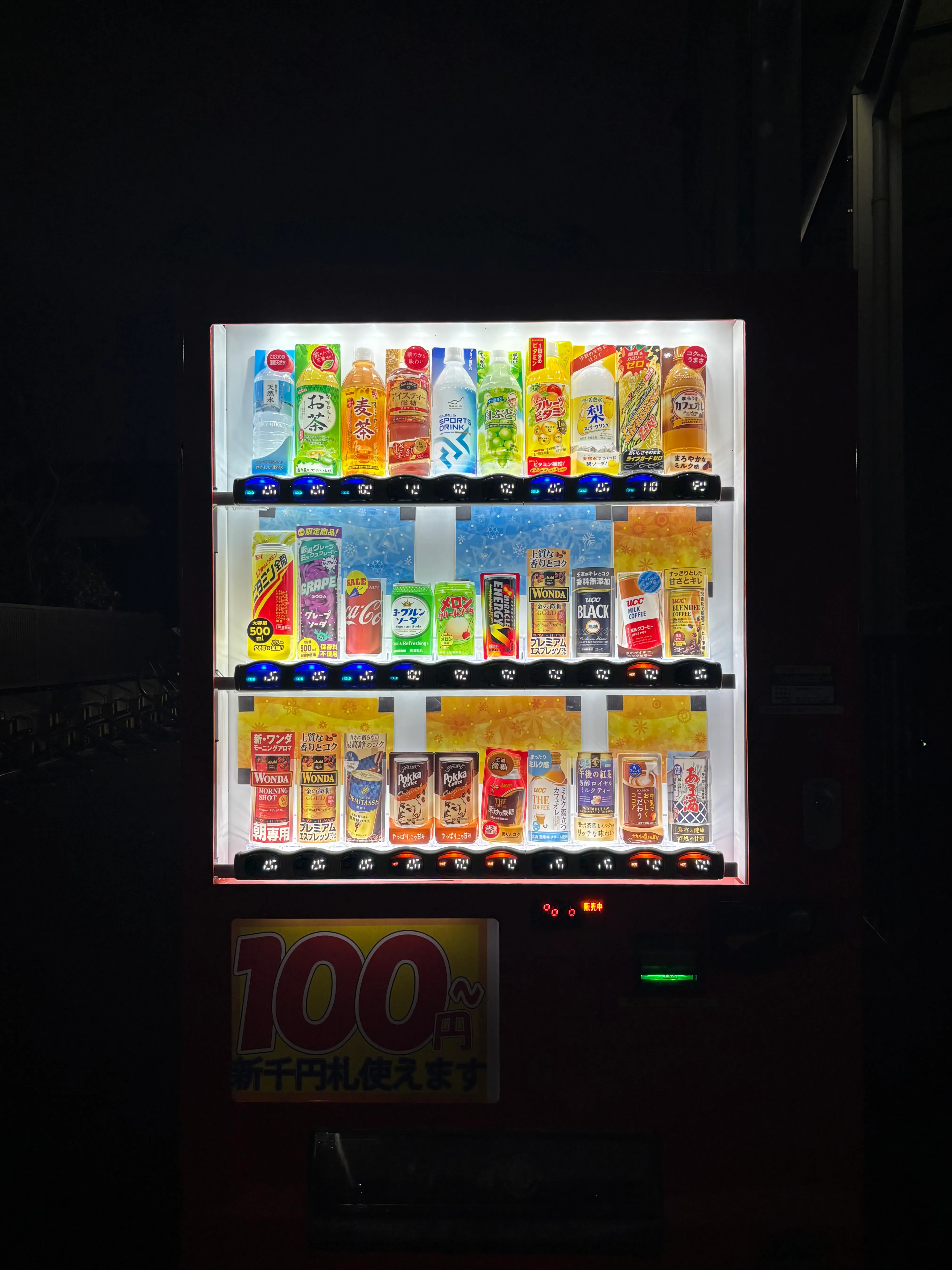 A Japanese vending machine at night displaying three shelves of colorful drinks including teas, sodas, and juices. The illuminated display shows prices in yen, with a 100 yen sign visible at the bottom