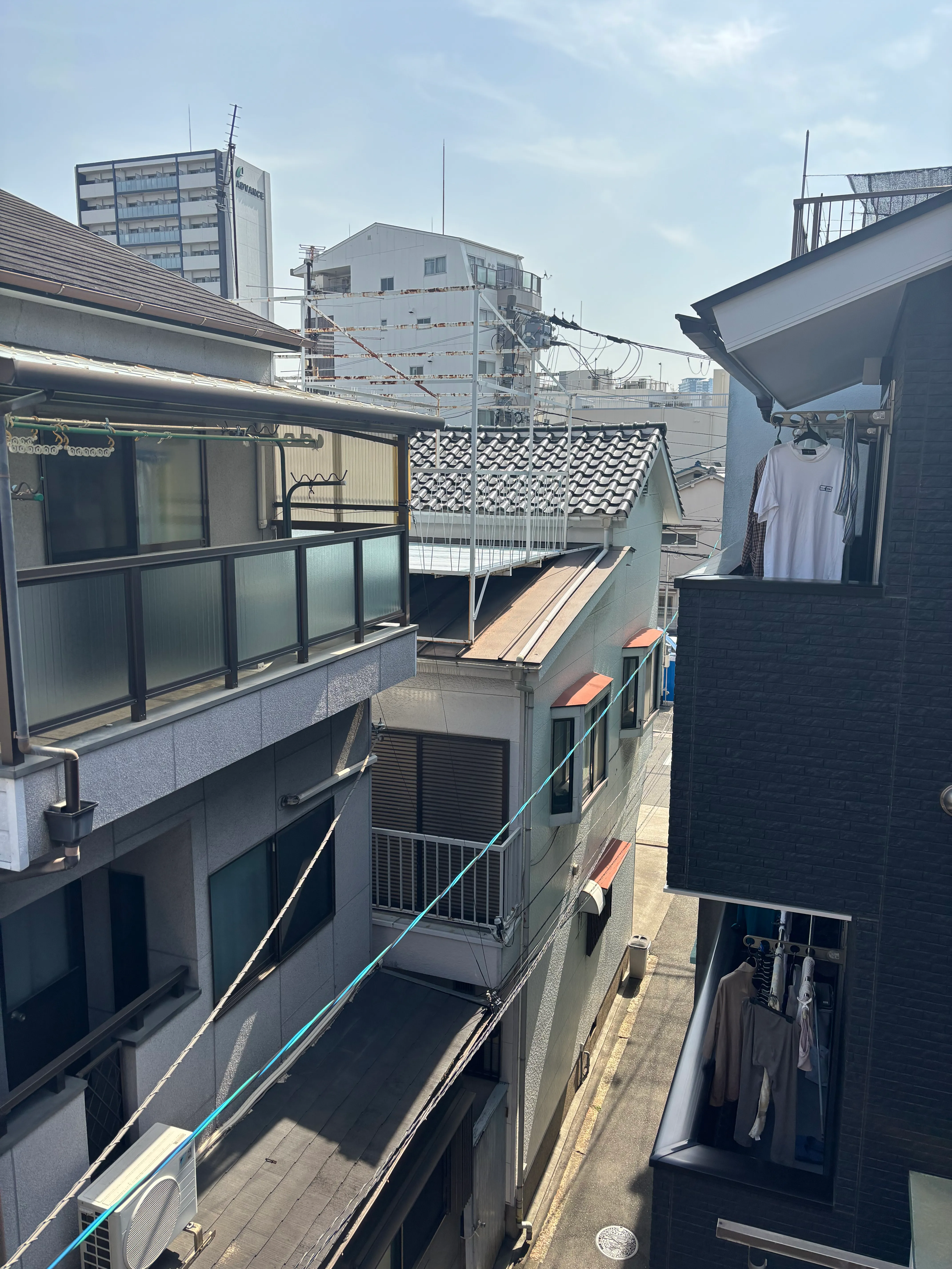 View looking down at densely packed Japanese residential buildings with various rooftop styles including traditional tile and modern flat roofs. Laundry lines stretch between buildings, with apartment towers visible in the background under a bright sky